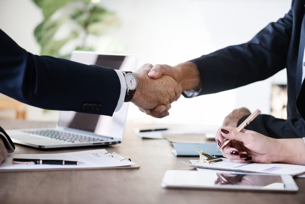 Poignée de main au dessus d'un bureau entre deux professionnels de l'immobilier, en arrière plan une personne est en train d'écrire. Sur la table sont posés un ordinateur portable et plusieurs papiers